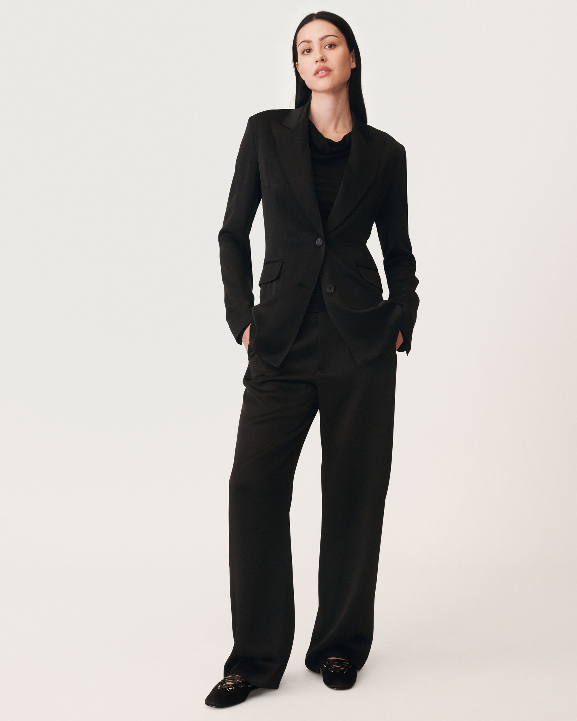 Woman standing against a white seamless background wearing a black blazer and matching trousers with her hands in her pockets