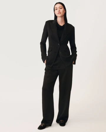 Woman standing against a white seamless background wearing a black blazer and matching trousers with her hands in her pockets