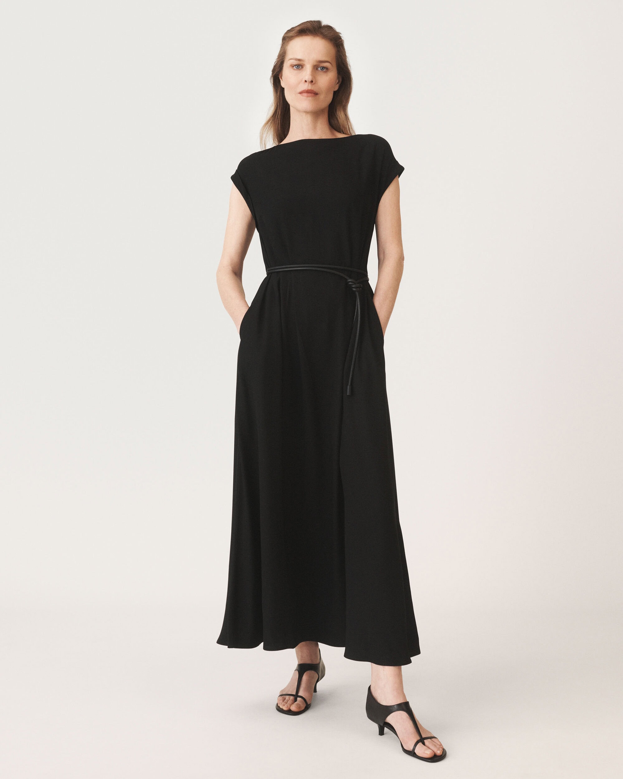 Model standing in a long black dress with a thin belt and sandals against a seamless white background