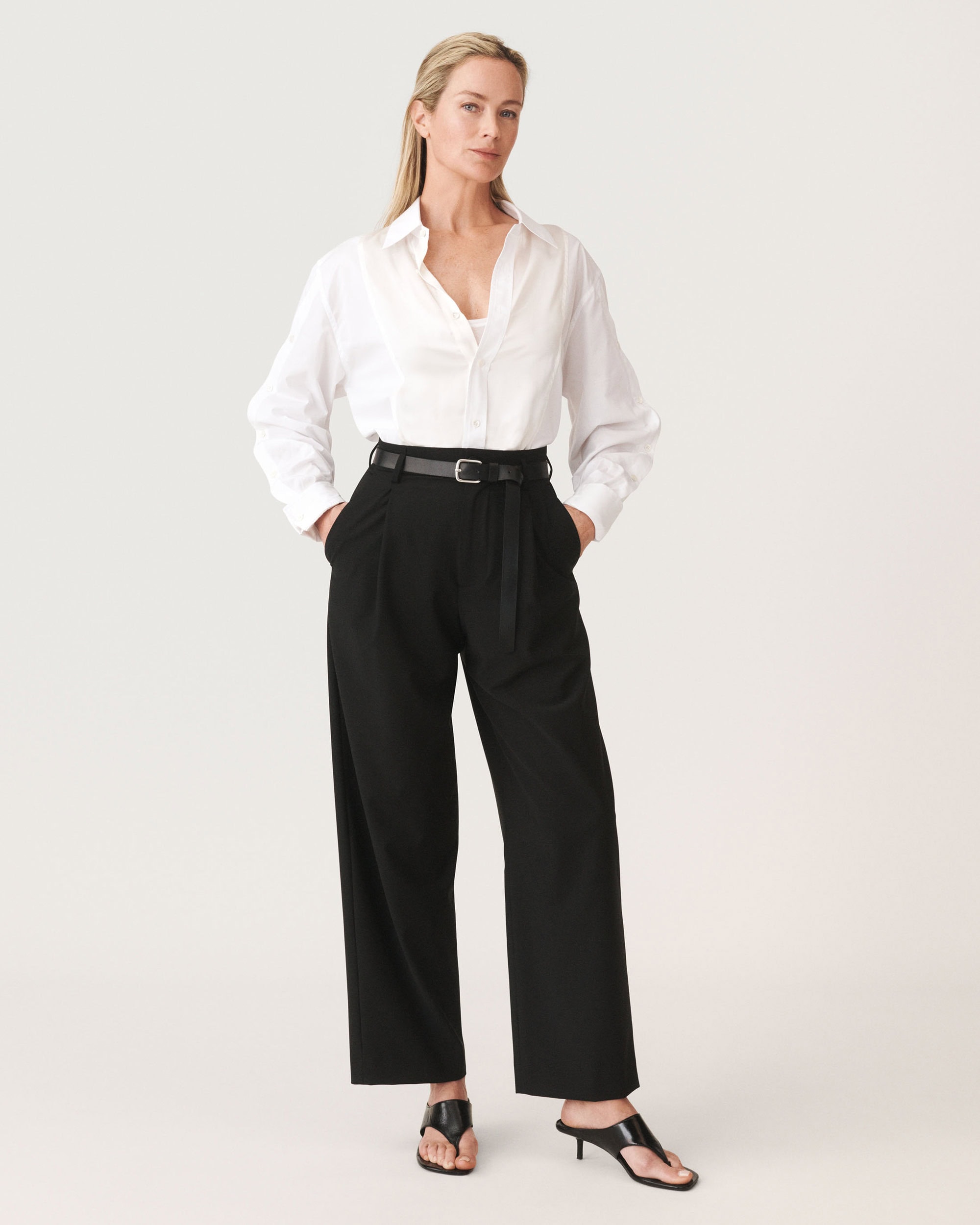 Full-length studio portrait of a woman wearing a white button-down shirt, black trousers, and a black belt against a clean white backdrop.