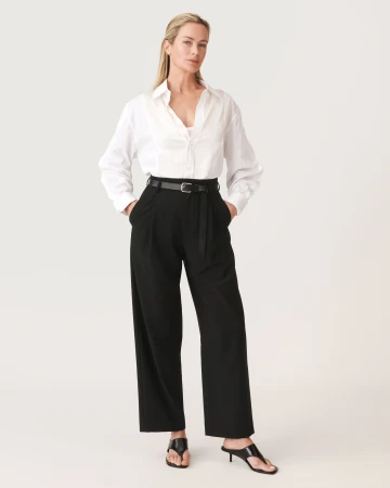 Full-length studio portrait of a woman wearing a white button-down shirt, black trousers, and a black belt against a clean white backdrop.
