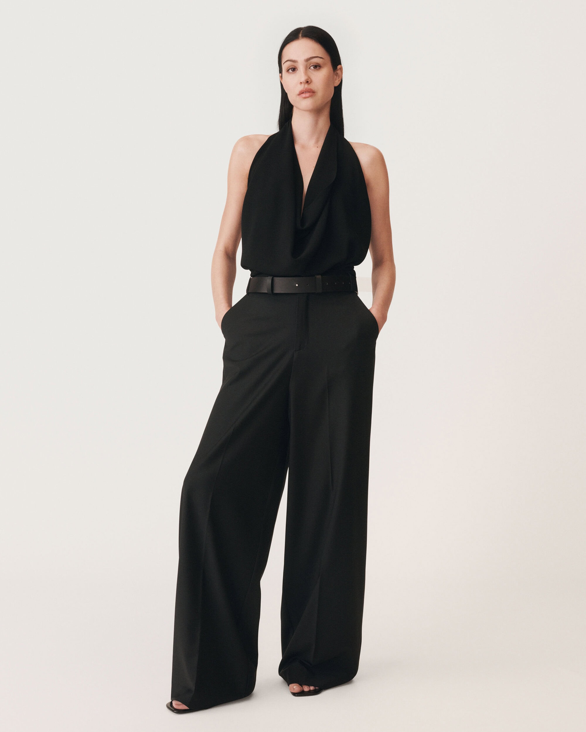 Model standing against a plain white background wearing a black halter top and black trousers with a belt