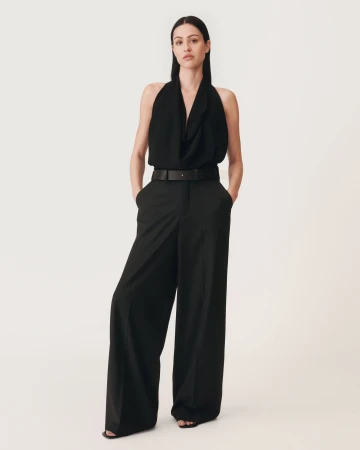 Model standing against a plain white background wearing a black halter top and black trousers with a belt
