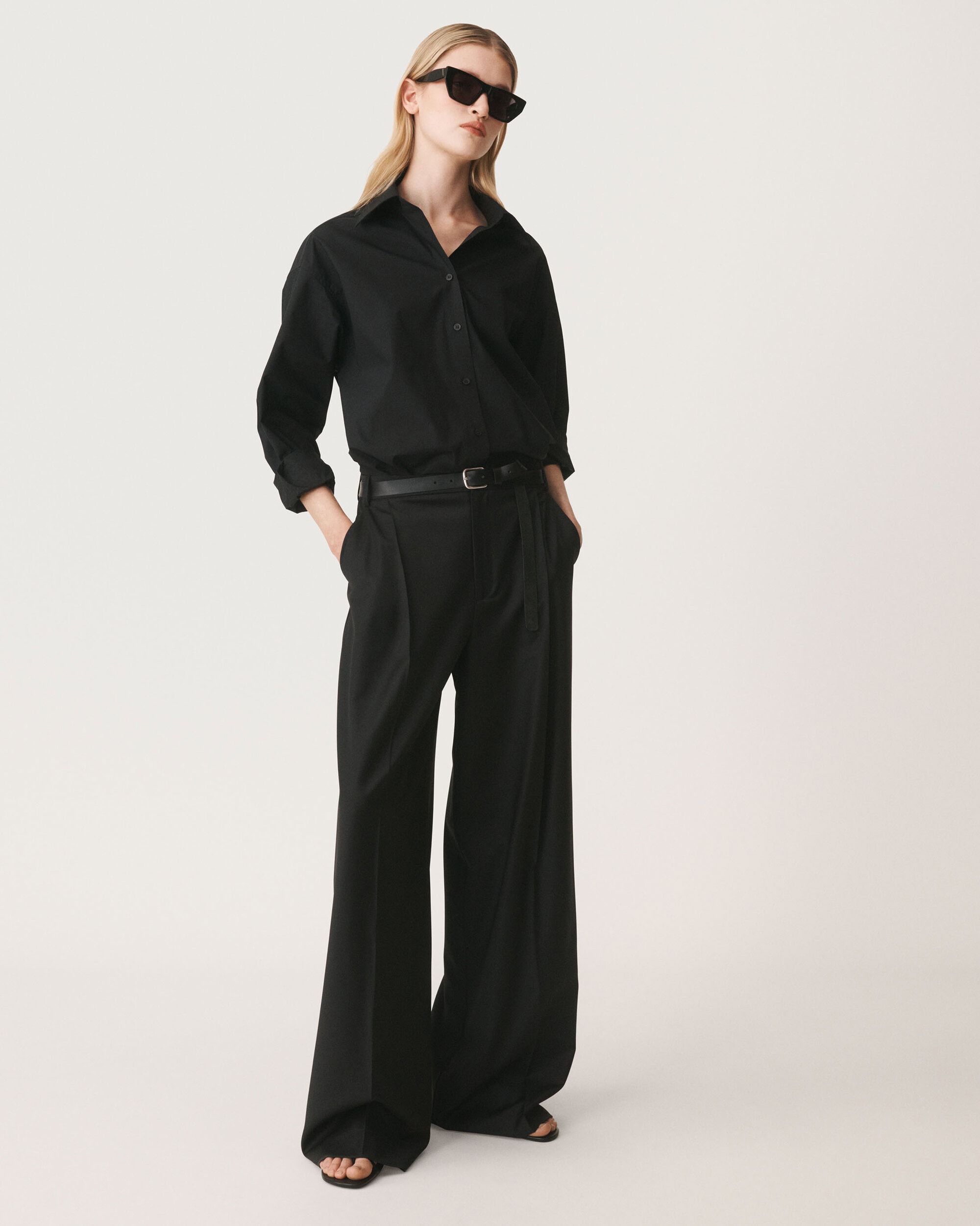 Model in black sunglasses wearing a black buttoned shirt and wide trousers against a white seamless background