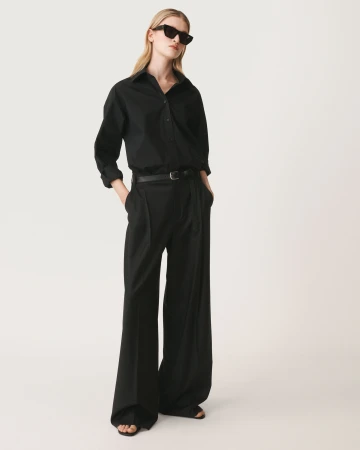 Model in black sunglasses wearing a black buttoned shirt and wide trousers against a white seamless background
