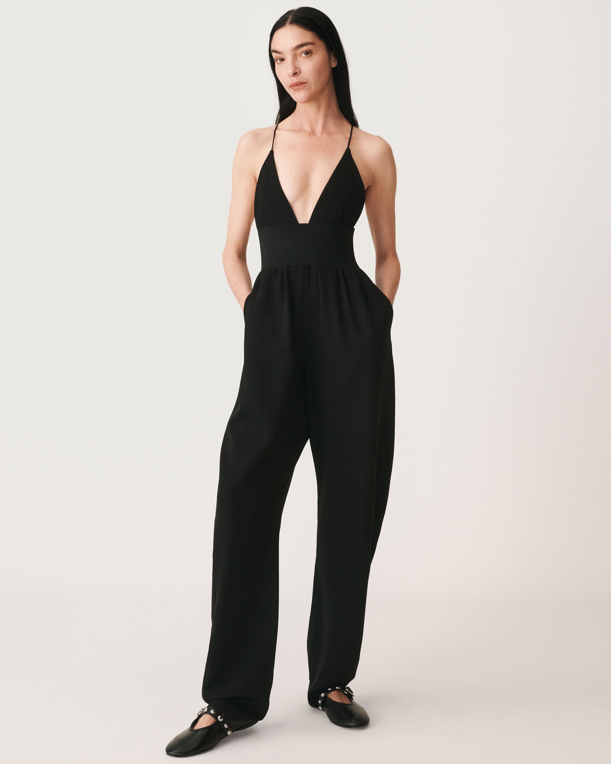 Model wearing a black jumpsuit with a deep V-neck and black flats against a seamless white background.