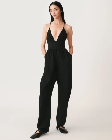 Model wearing a black jumpsuit with a deep V-neck and black flats against a seamless white background.