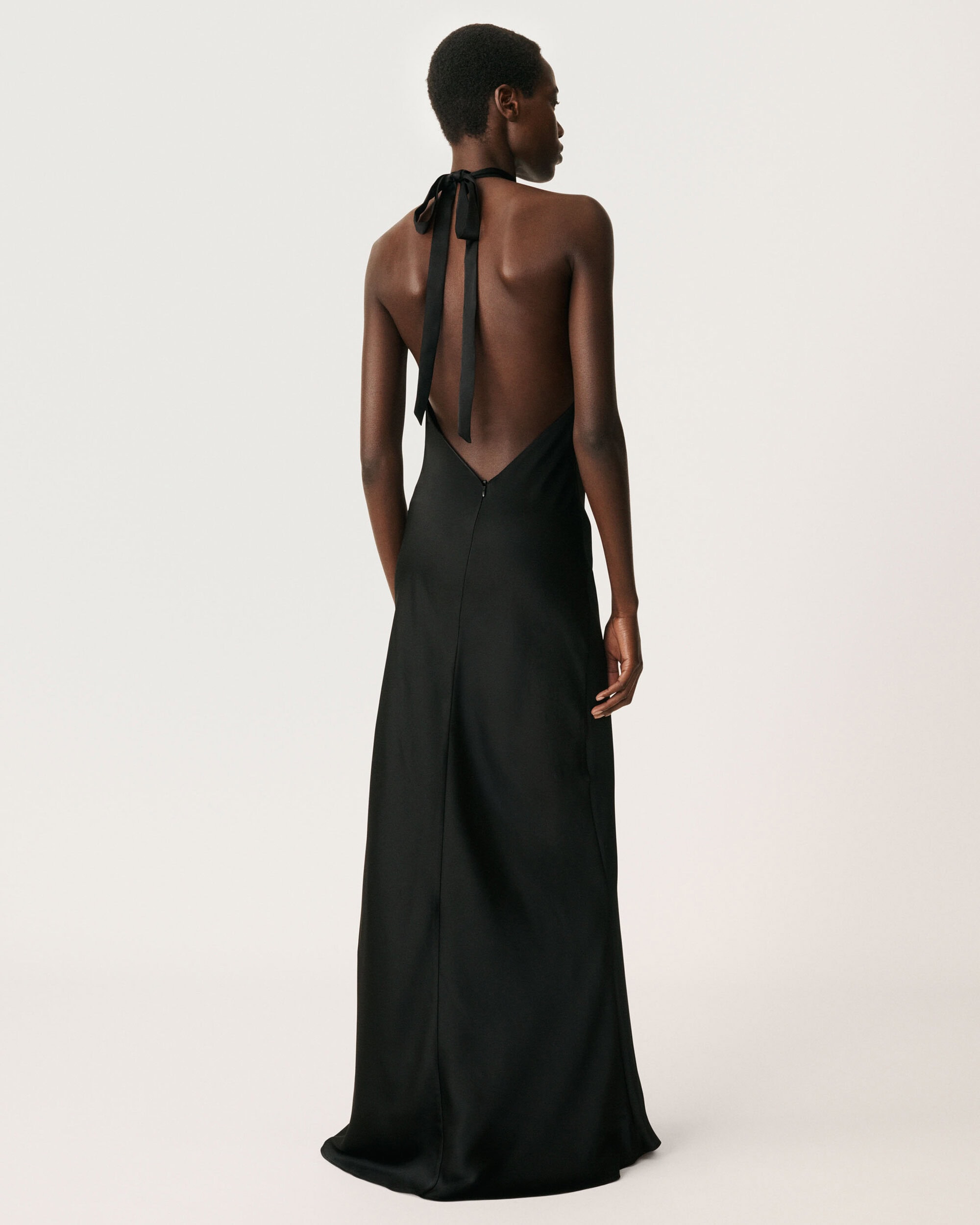Black woman with short dark hair viewed from behind, wearing a long black halter-neck slip dress against a plain white seamless backdrop.