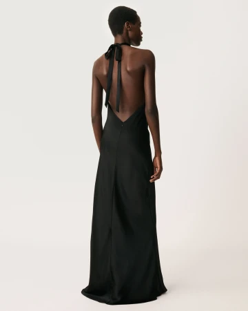 Black woman with short dark hair viewed from behind, wearing a long black halter-neck slip dress against a plain white seamless backdrop.