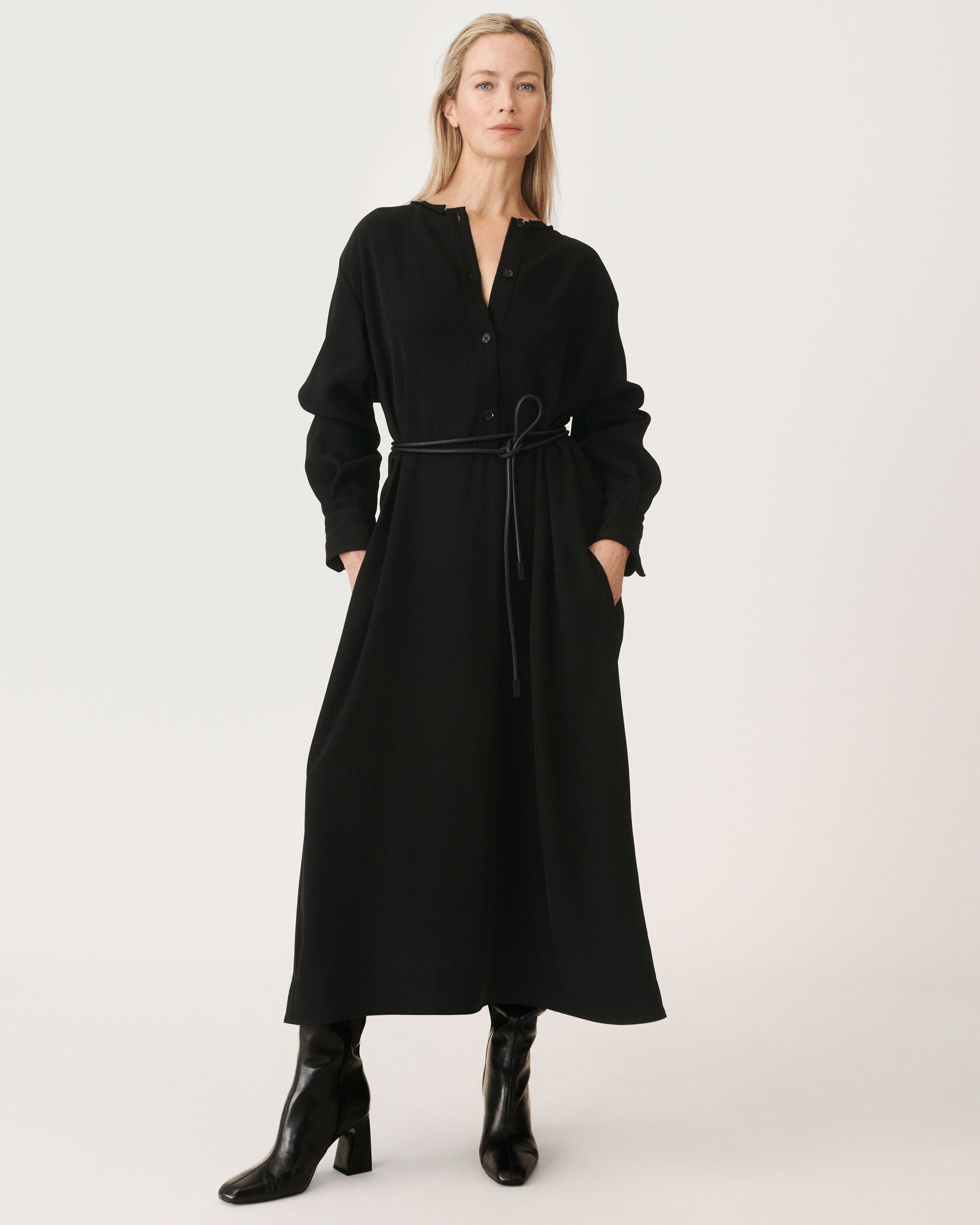 Carolyn Murphy in a long black belted dress and black boots against a plain white seamless studio background.