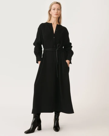Carolyn Murphy in a long black belted dress and black boots against a plain white seamless studio background.