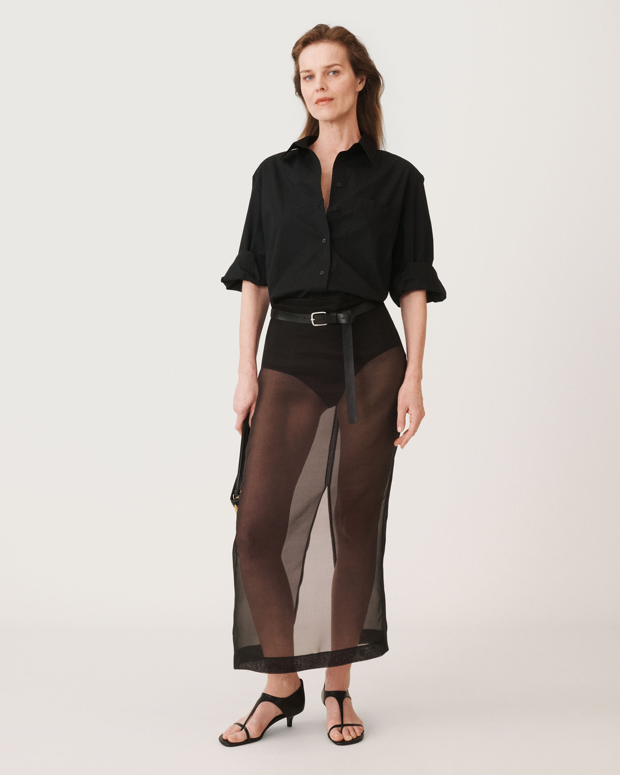 Model standing against a plain white background wearing a black buttoned shirt and sheer skirt with a thin belt.