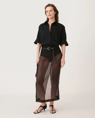 Model standing against a plain white background wearing a black buttoned shirt and sheer skirt with a thin belt.