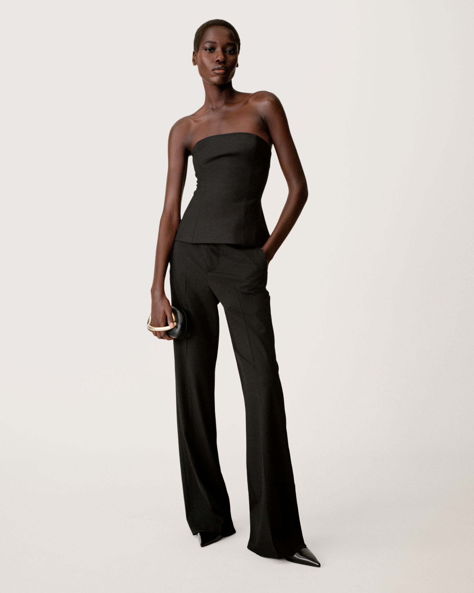 Model in a black strapless top and trousers holding a round black handbag against a light seamless background