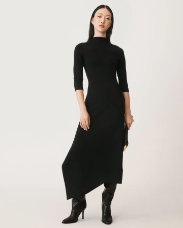 Model standing against a plain white backdrop wearing a black long-sleeved dress and black boots, holding a clutch.