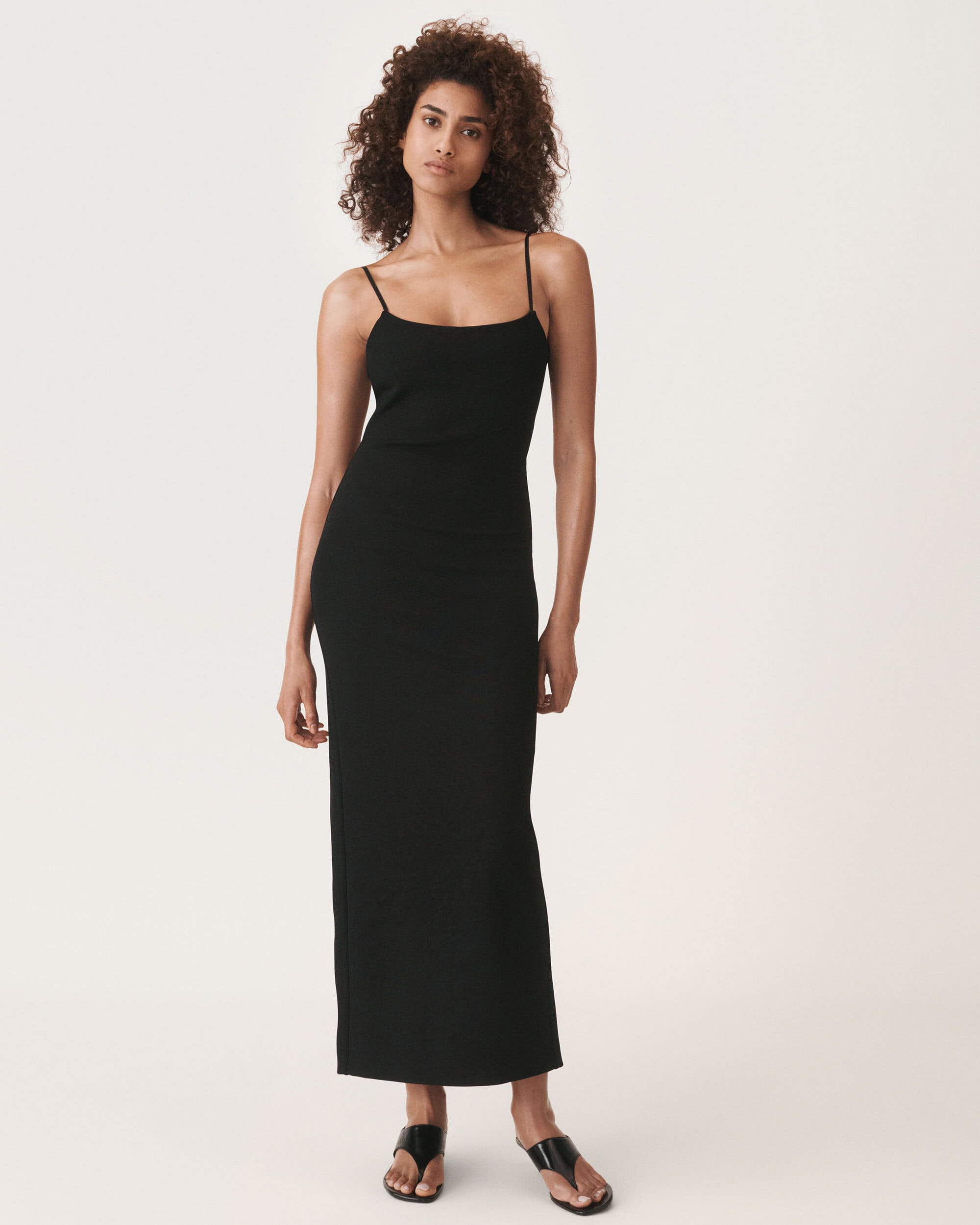 Model wearing a black slip dress and sandals standing against a plain white background for Zara.