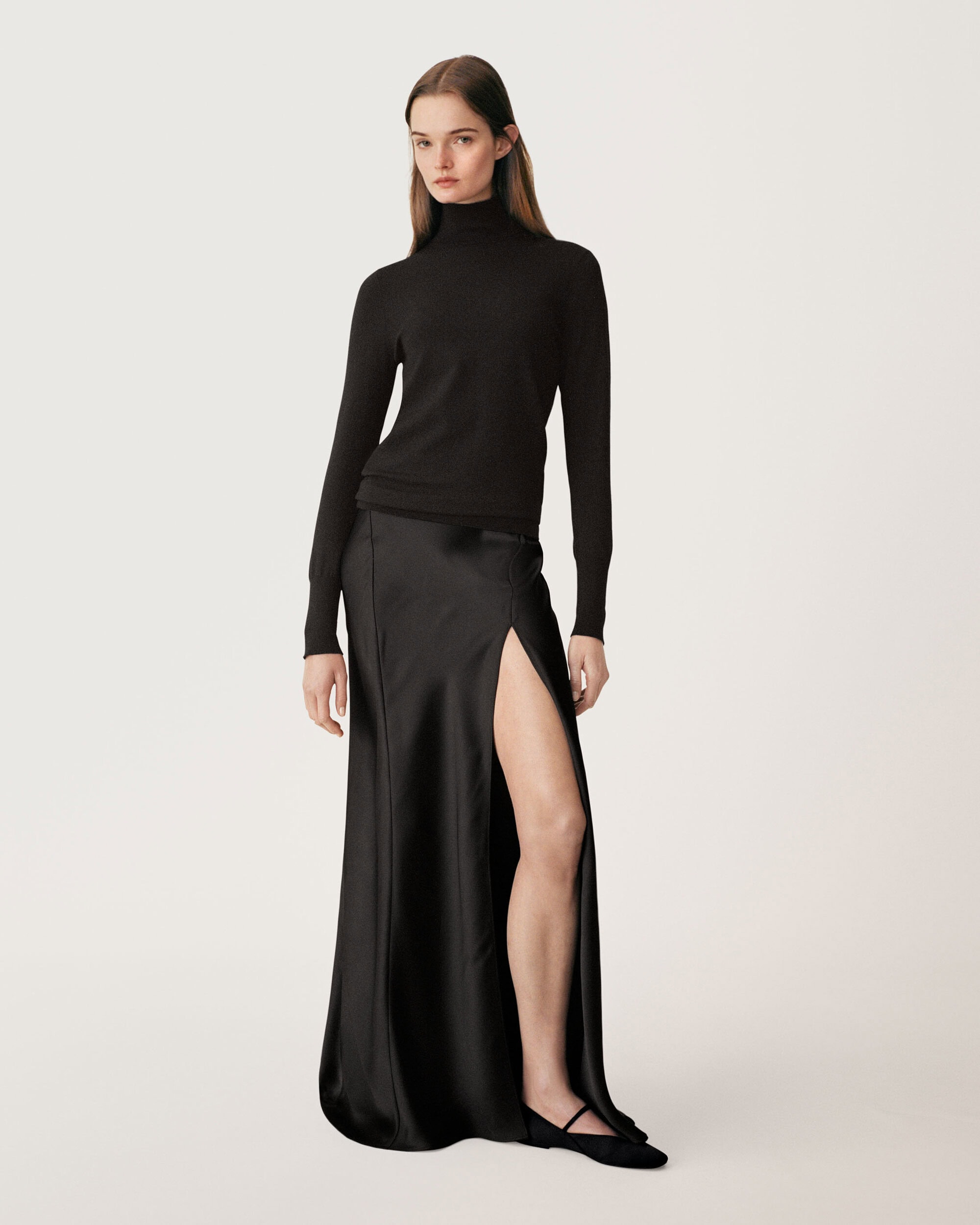 Full-length fashion portrait of a female model wearing a black turtleneck and long black skirt with a high slit against a white background