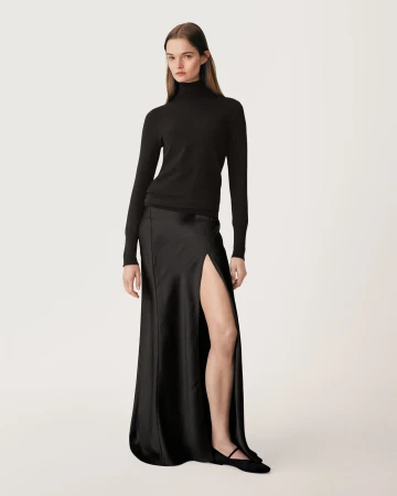 Full-length fashion portrait of a female model wearing a black turtleneck and long black skirt with a high slit against a white background
