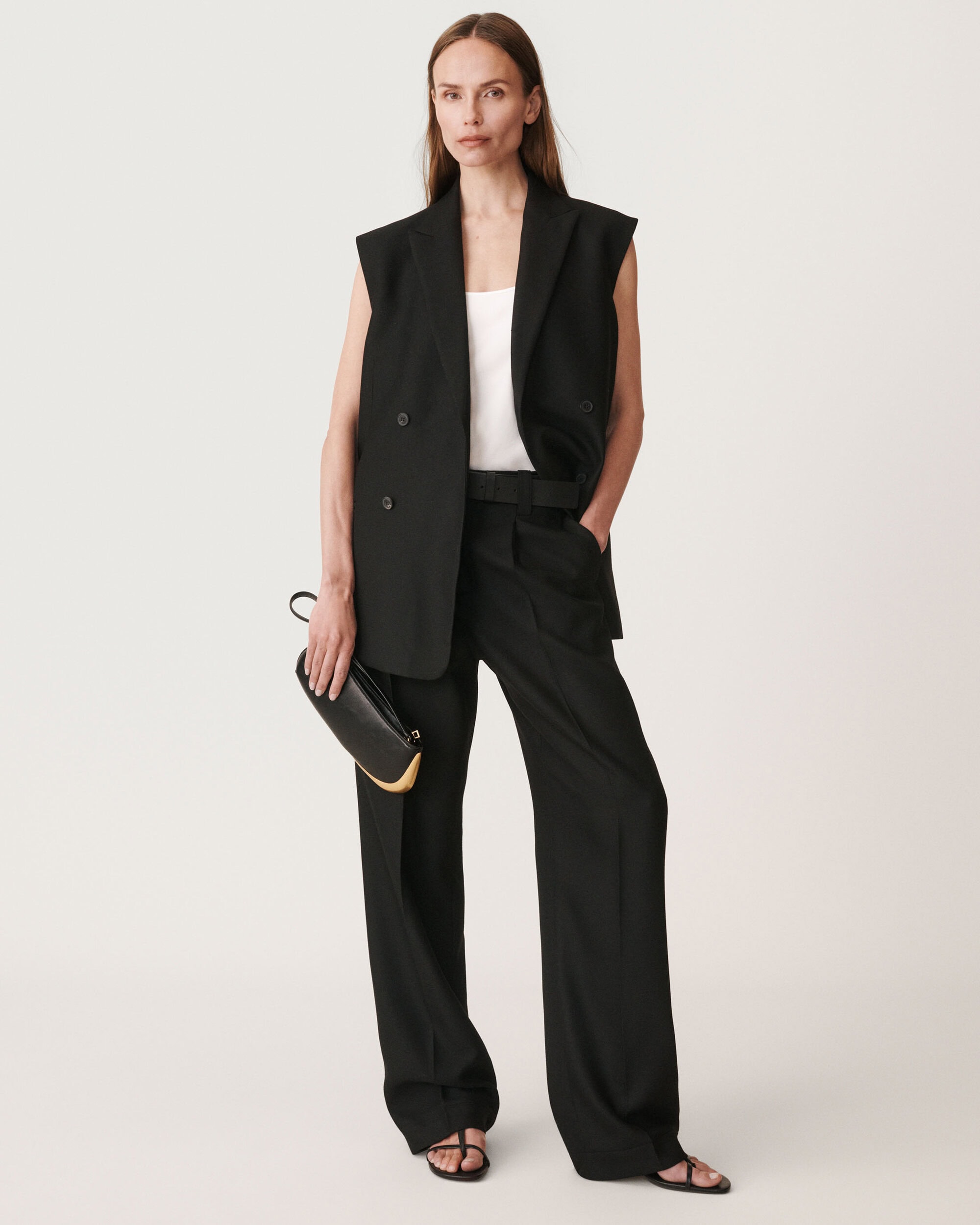 Model wearing a black vest and trousers holding a black clutch against a plain white background