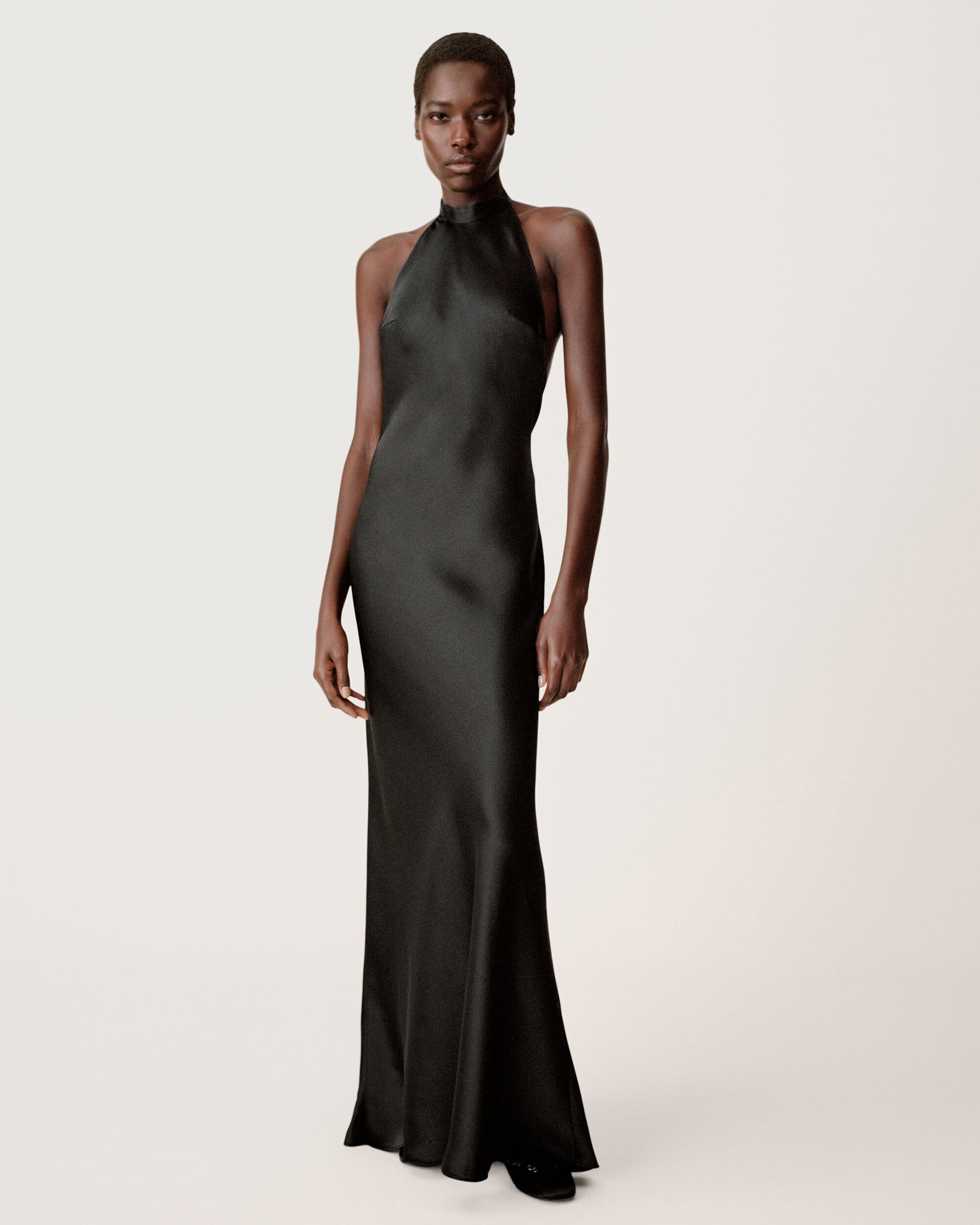 Black woman with short hair wearing a long black halter-neck evening dress against a plain neutral background.