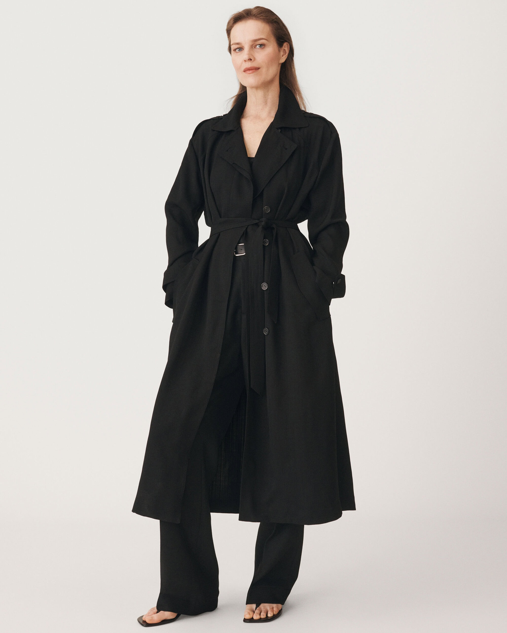 Model wearing a long black belted coat and dark trousers standing against a plain white background