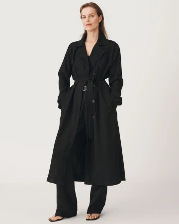 Model wearing a long black belted coat and dark trousers standing against a plain white background