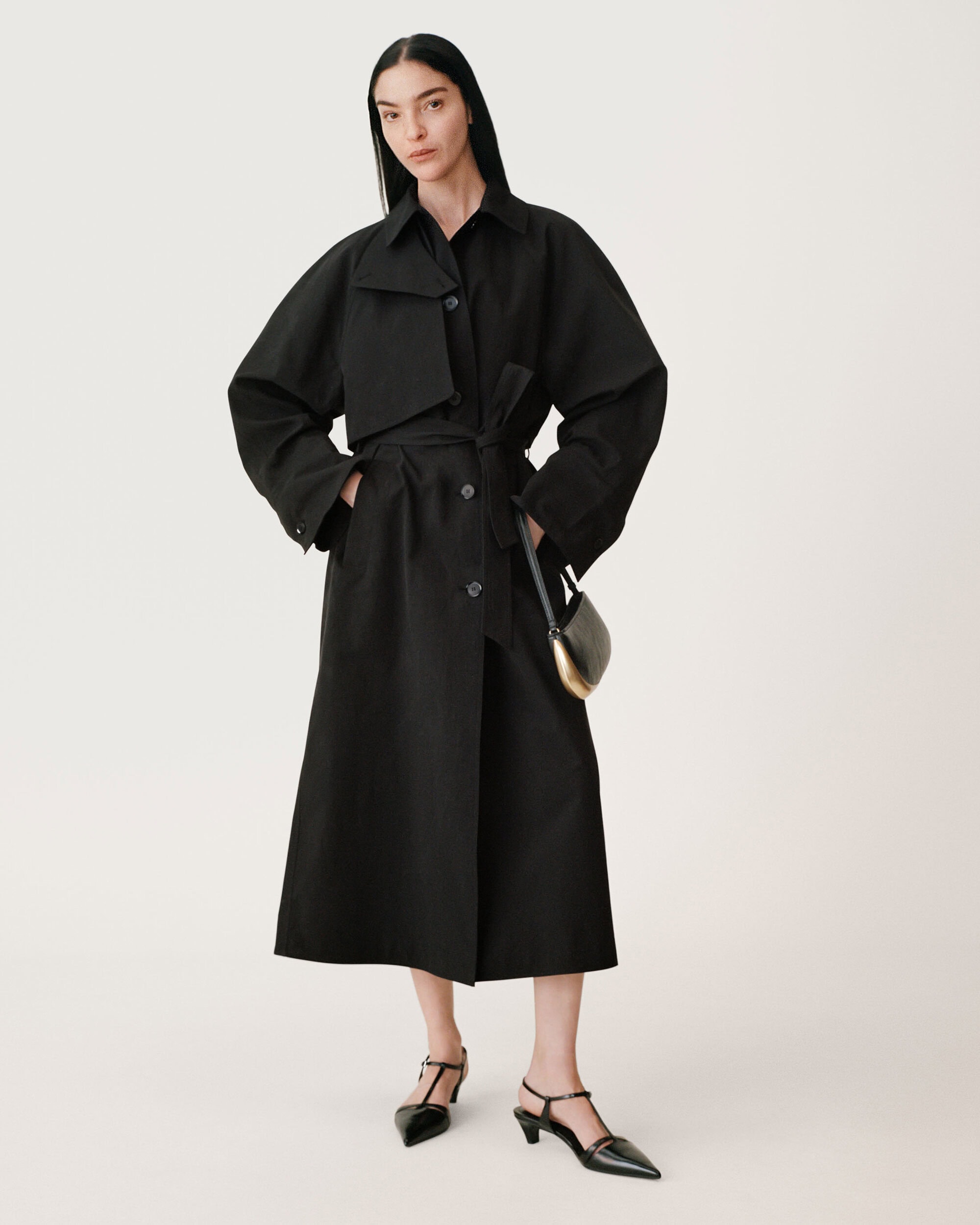 Model wearing a long black belted coat with dark sunglasses against a seamless white background for Zara