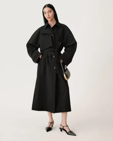 Model wearing a long black belted coat with dark sunglasses against a seamless white background for Zara