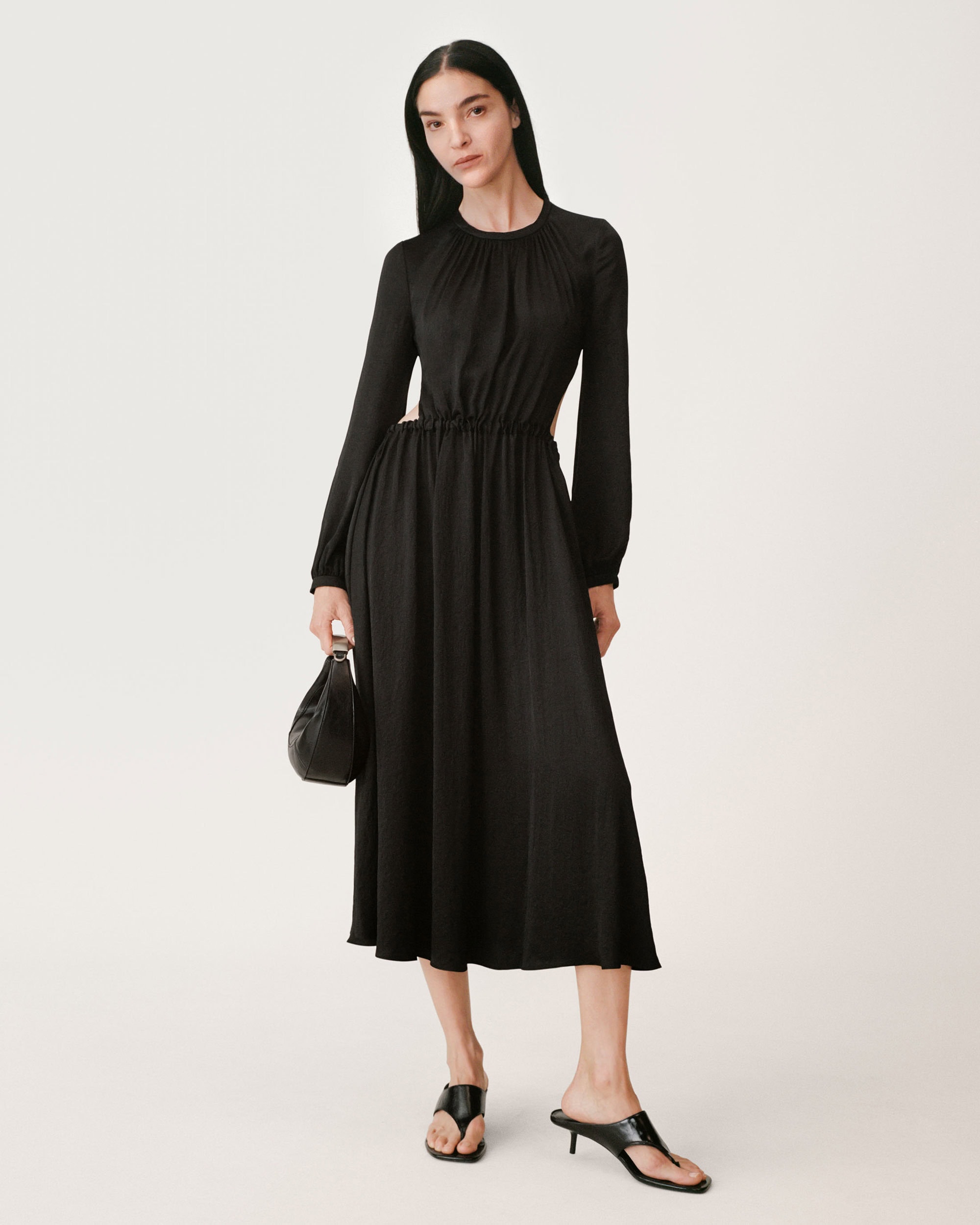Model in long black gathered dress holding a black handbag against a white background