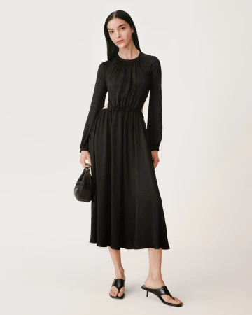 Model in long black gathered dress holding a black handbag against a white background