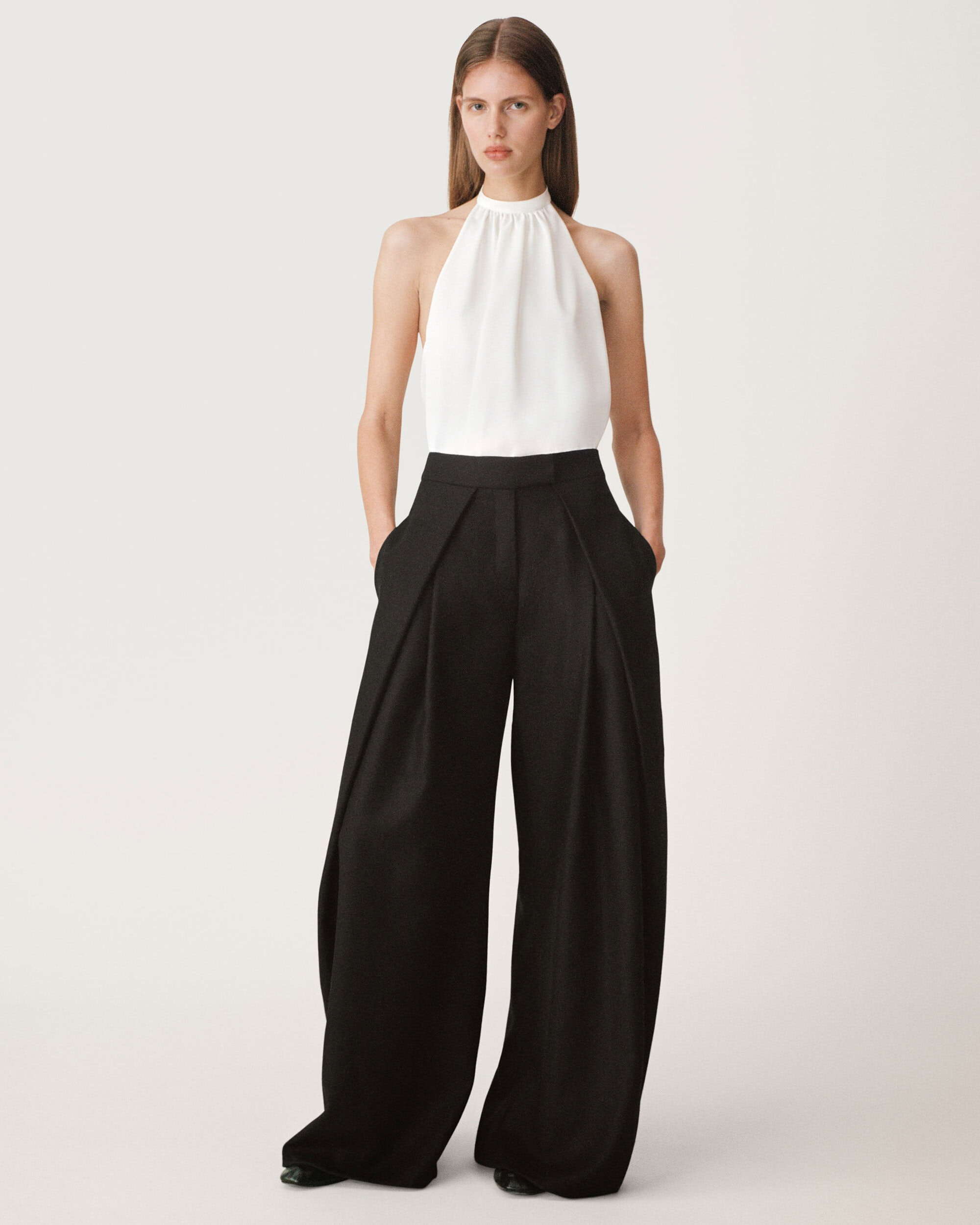 A female model standing against a white seamless background wearing a white halter top and high waisted black wide leg trousers.