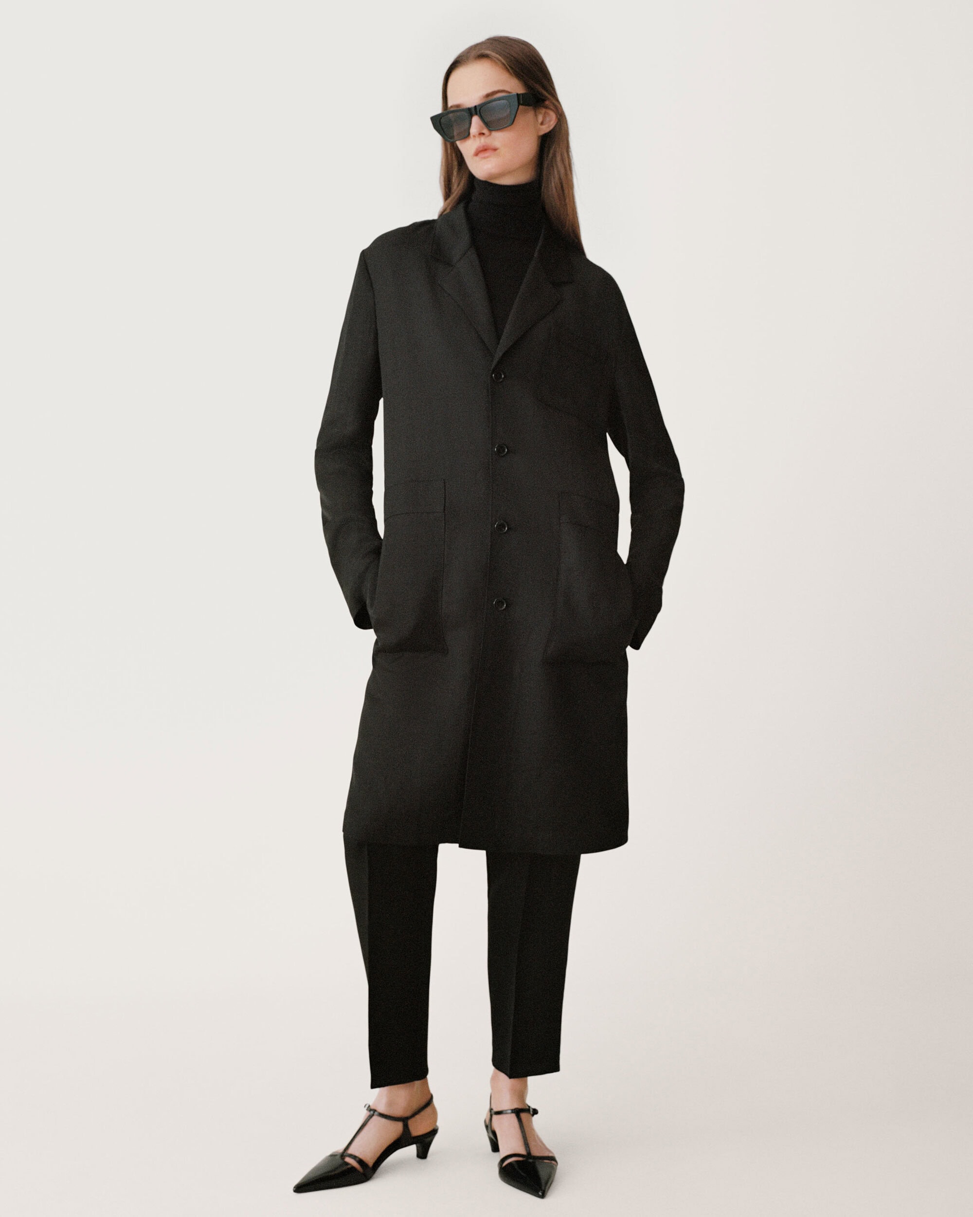 Model in dark sunglasses wearing a long black coat against a neutral white background