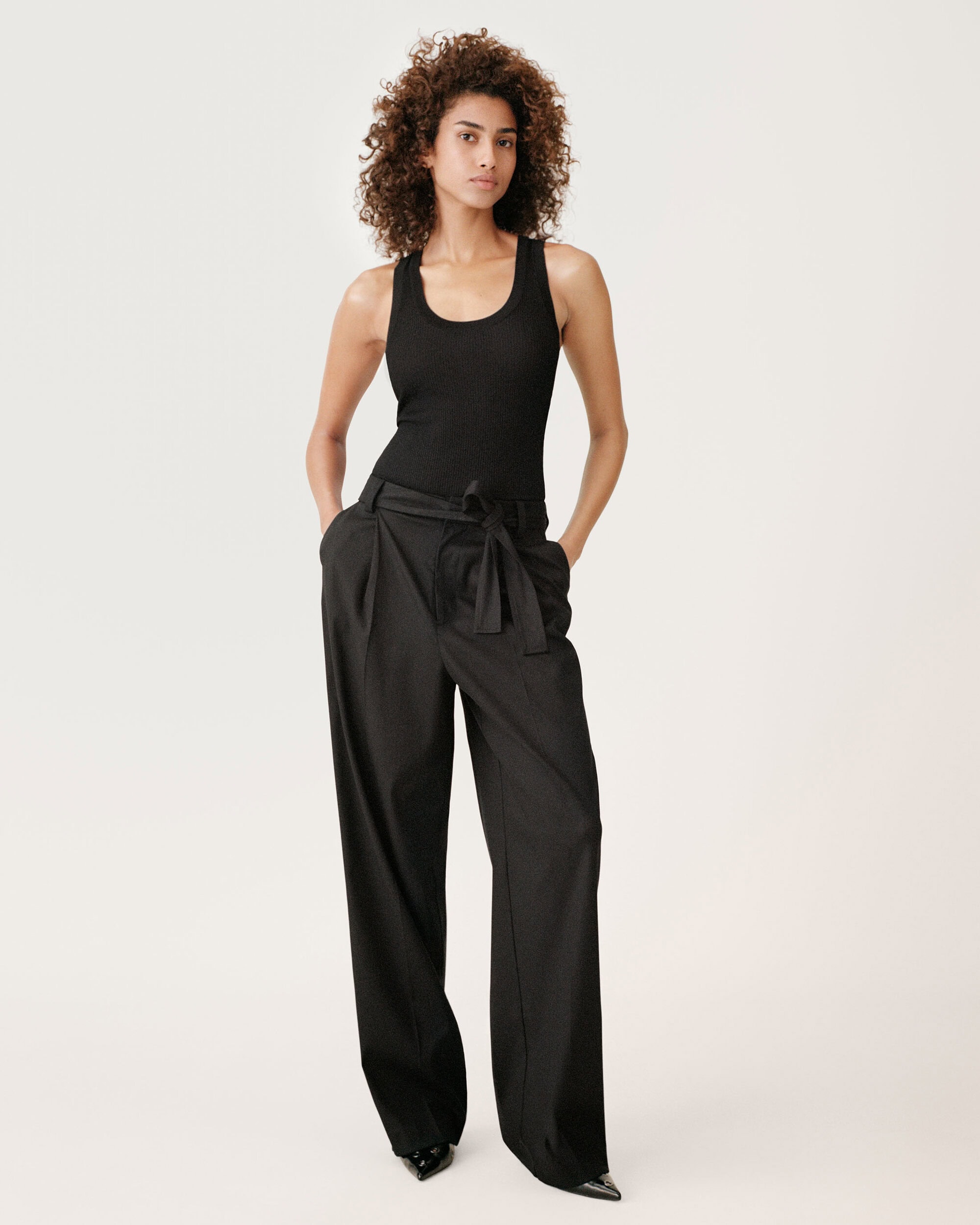 Woman with curly dark hair wearing a black tank top and wide trousers standing against a white background.