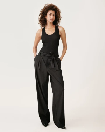 Woman with curly dark hair wearing a black tank top and wide trousers standing against a white background.