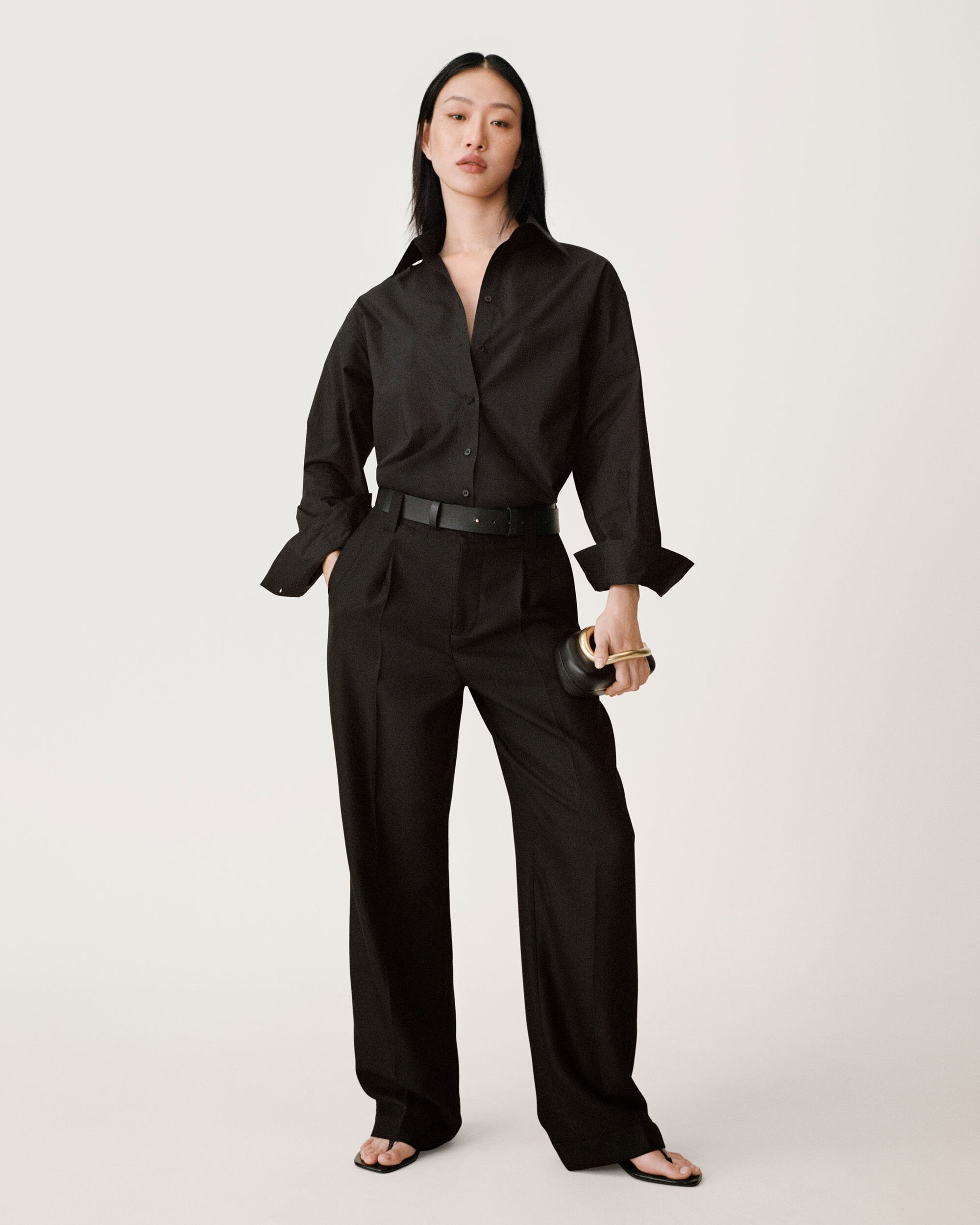 Asian woman in black button-down shirt and black trousers holding a small black handbag against a seamless white background.