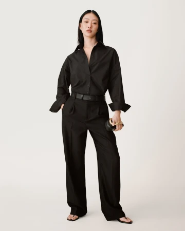 Asian woman in black button-down shirt and black trousers holding a small black handbag against a seamless white background.