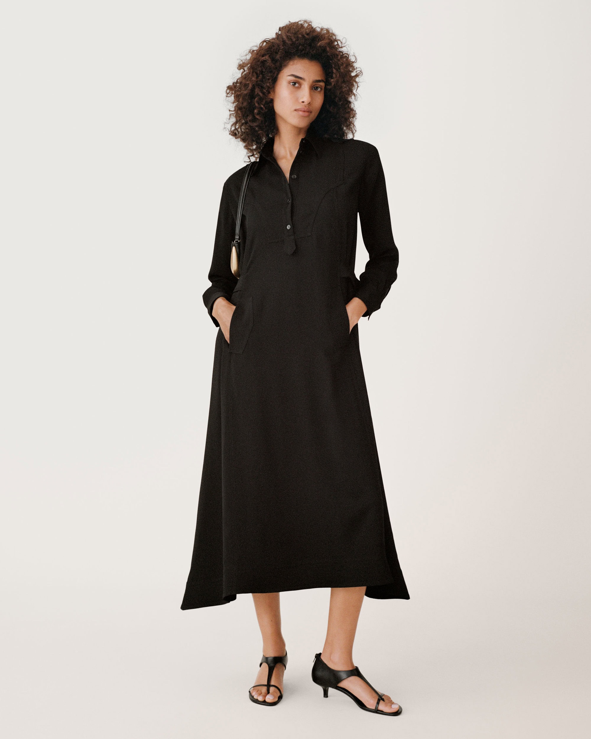 Model wearing a long black button-down dress and black sandals against a solid white background