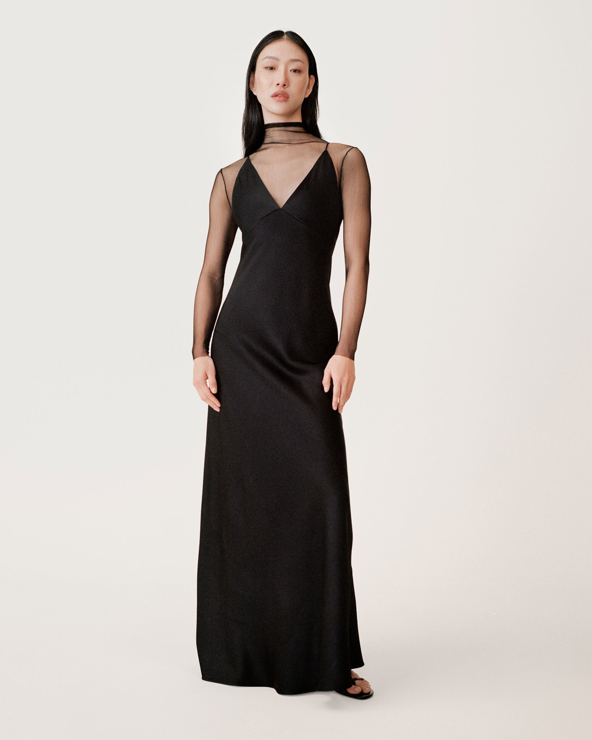 Model in black slip dress with sheer long sleeves standing against a seamless white studio background