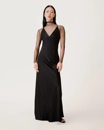 Model in black slip dress with sheer long sleeves standing against a seamless white studio background