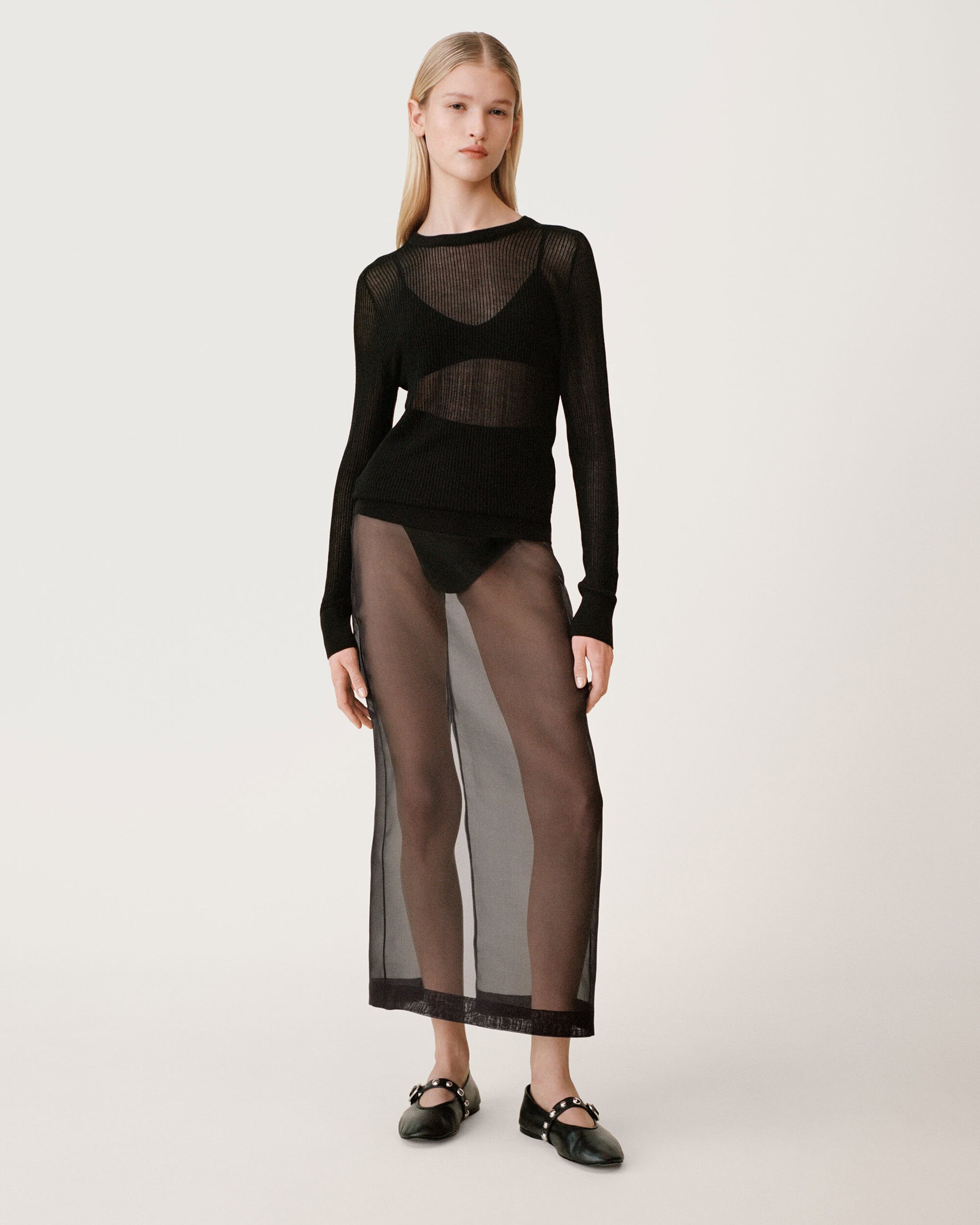 Model standing on white studio floor wearing a sheer black knit top and long translucent skirt with black flats