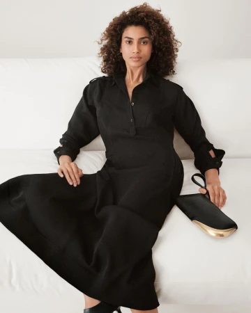 Model with curly hair seated on a white sofa wearing a long black dress and holding a black clutch bag