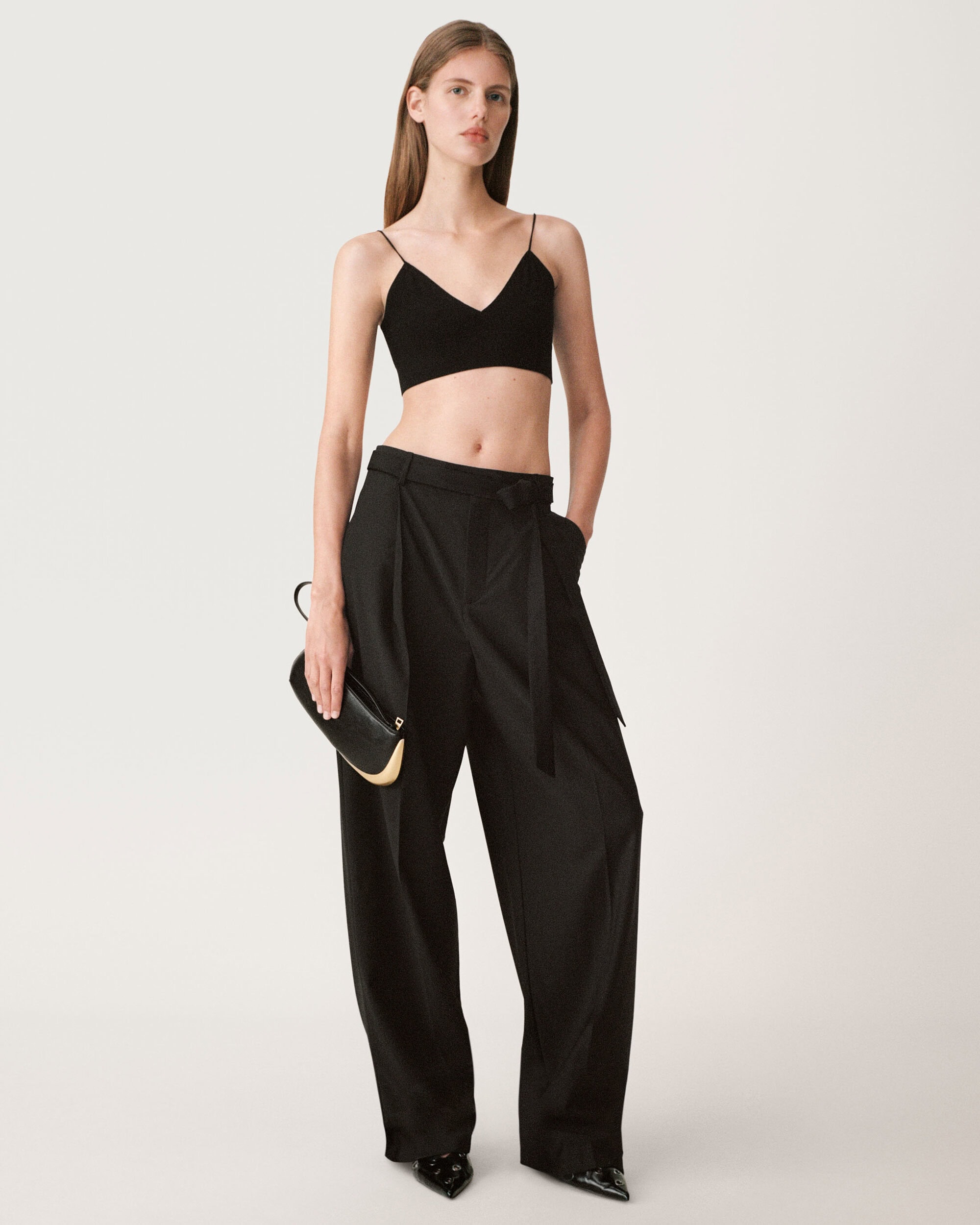Model wearing a black crop top and wide-leg trousers holding a clutch against a white seamless background