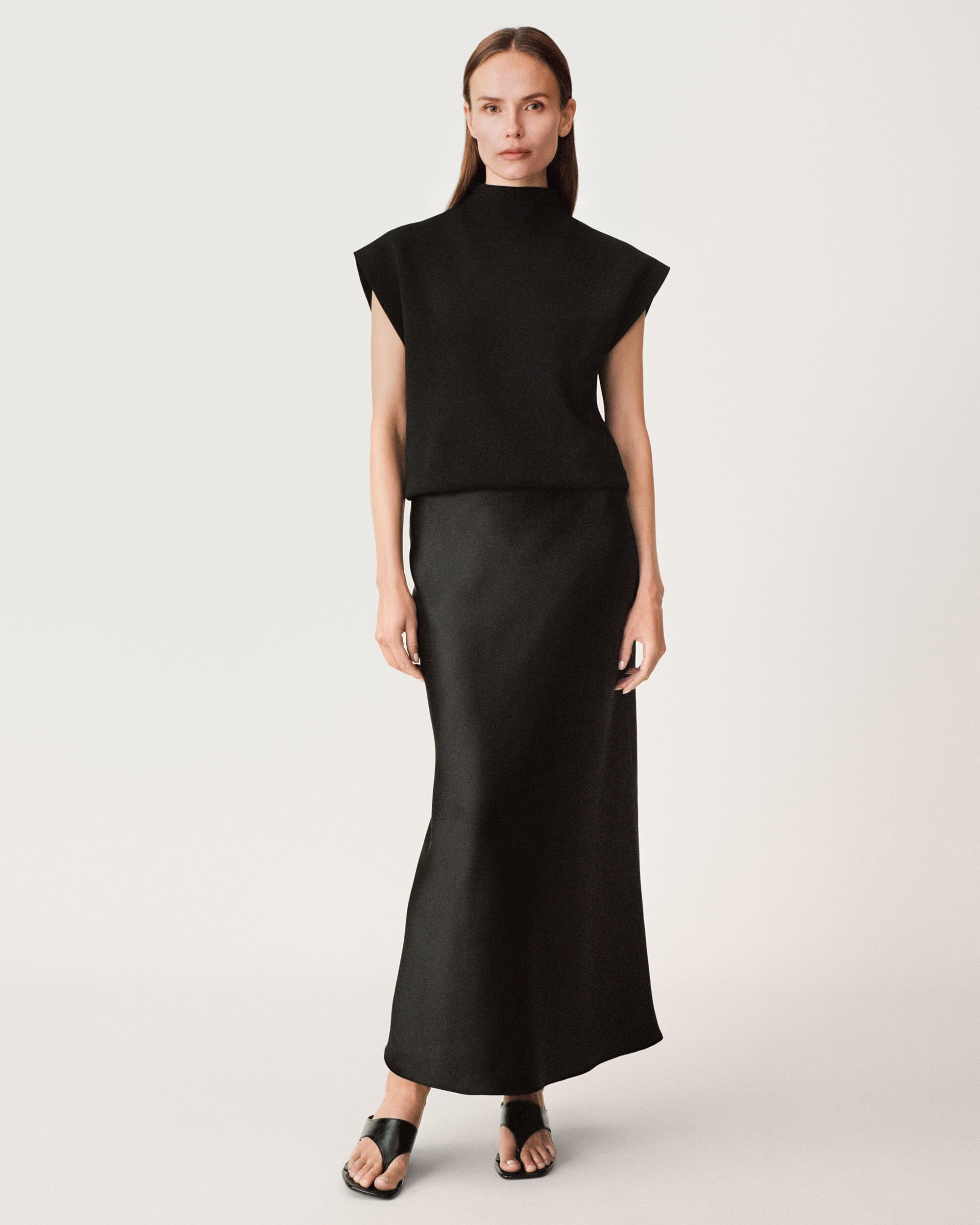 Model Amber Valletta wearing a black sleeveless top and long skirt against white seamless background