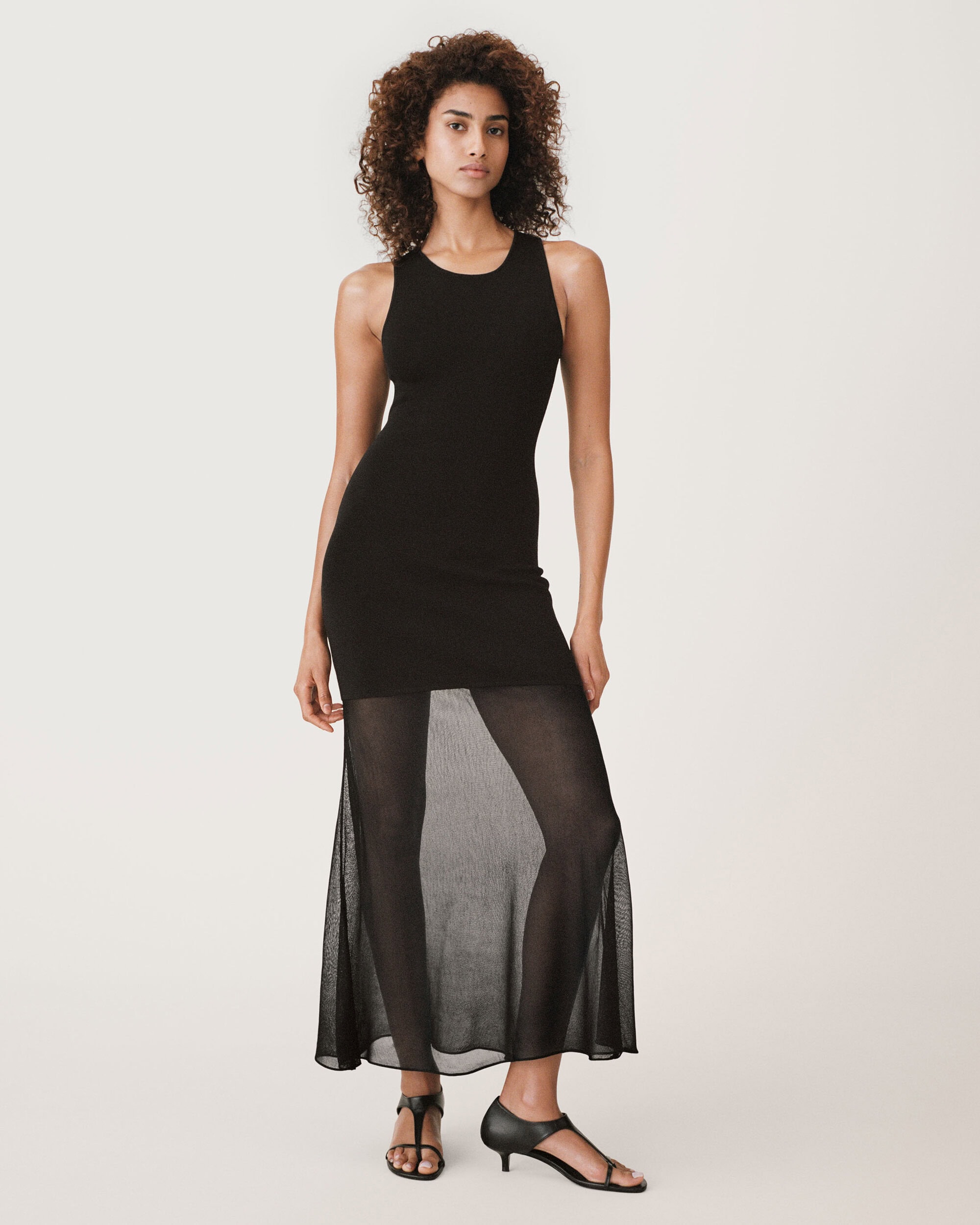 Model standing against a plain white backdrop wearing a black sleeveless midi dress with a sheer lower skirt and black strappy low heels.