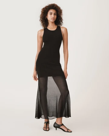 Model standing against a plain white backdrop wearing a black sleeveless midi dress with a sheer lower skirt and black strappy low heels.