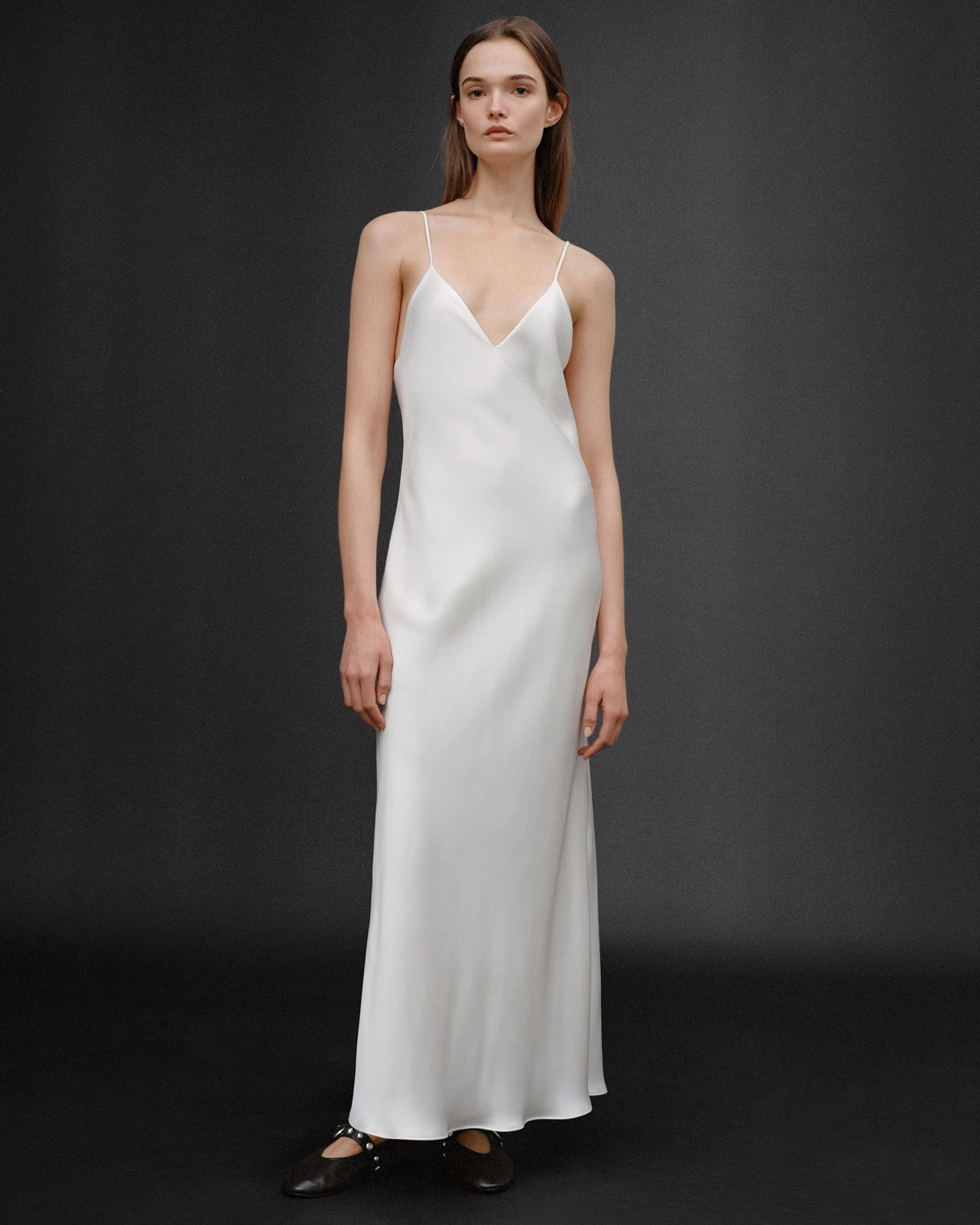 Model wearing a long white slip dress against a dark grey seamless studio backdrop