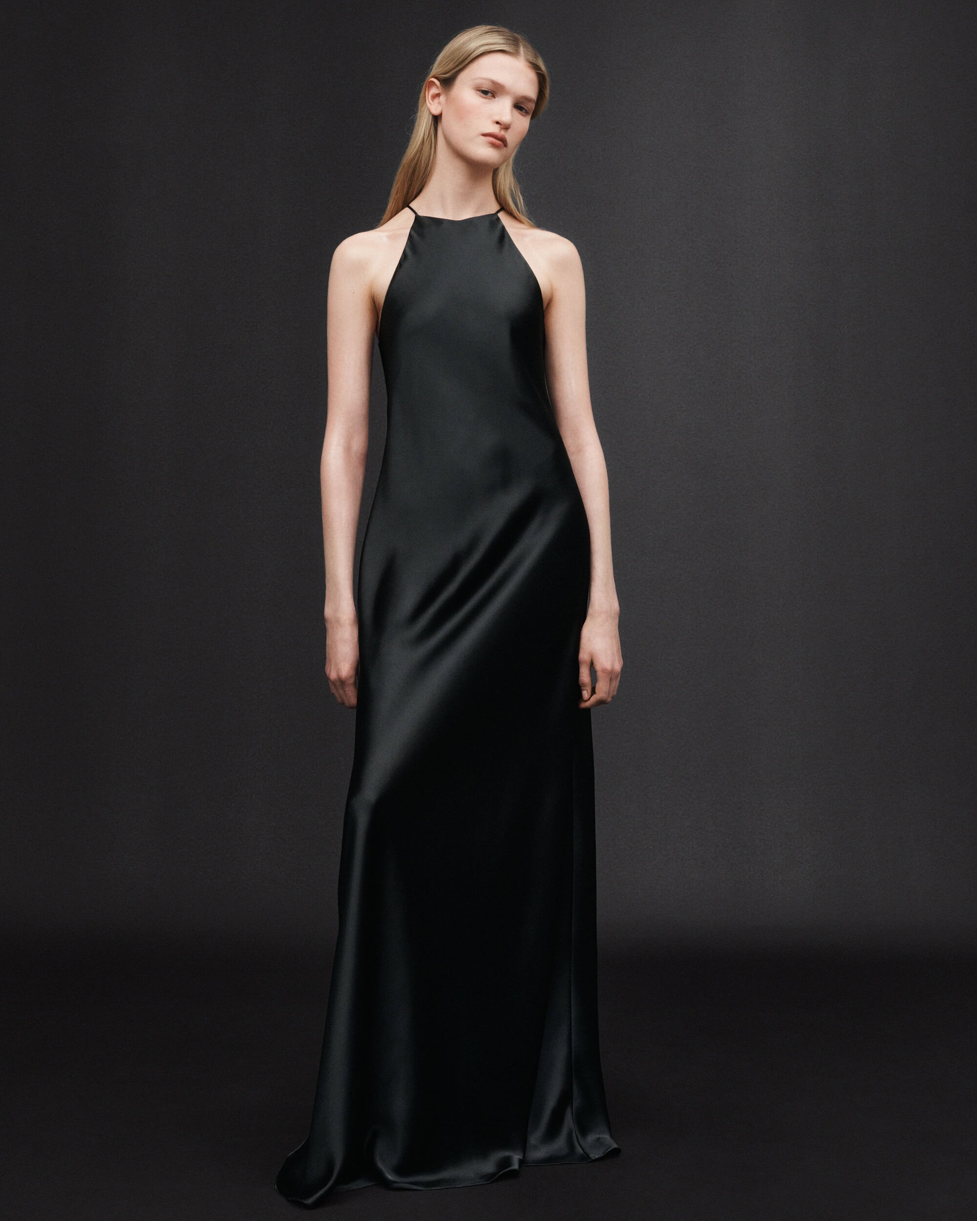 Model in a long black halter slip dress standing against a dark grey seamless studio backdrop.