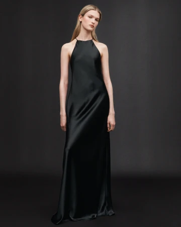 Model in a long black halter slip dress standing against a dark grey seamless studio backdrop.