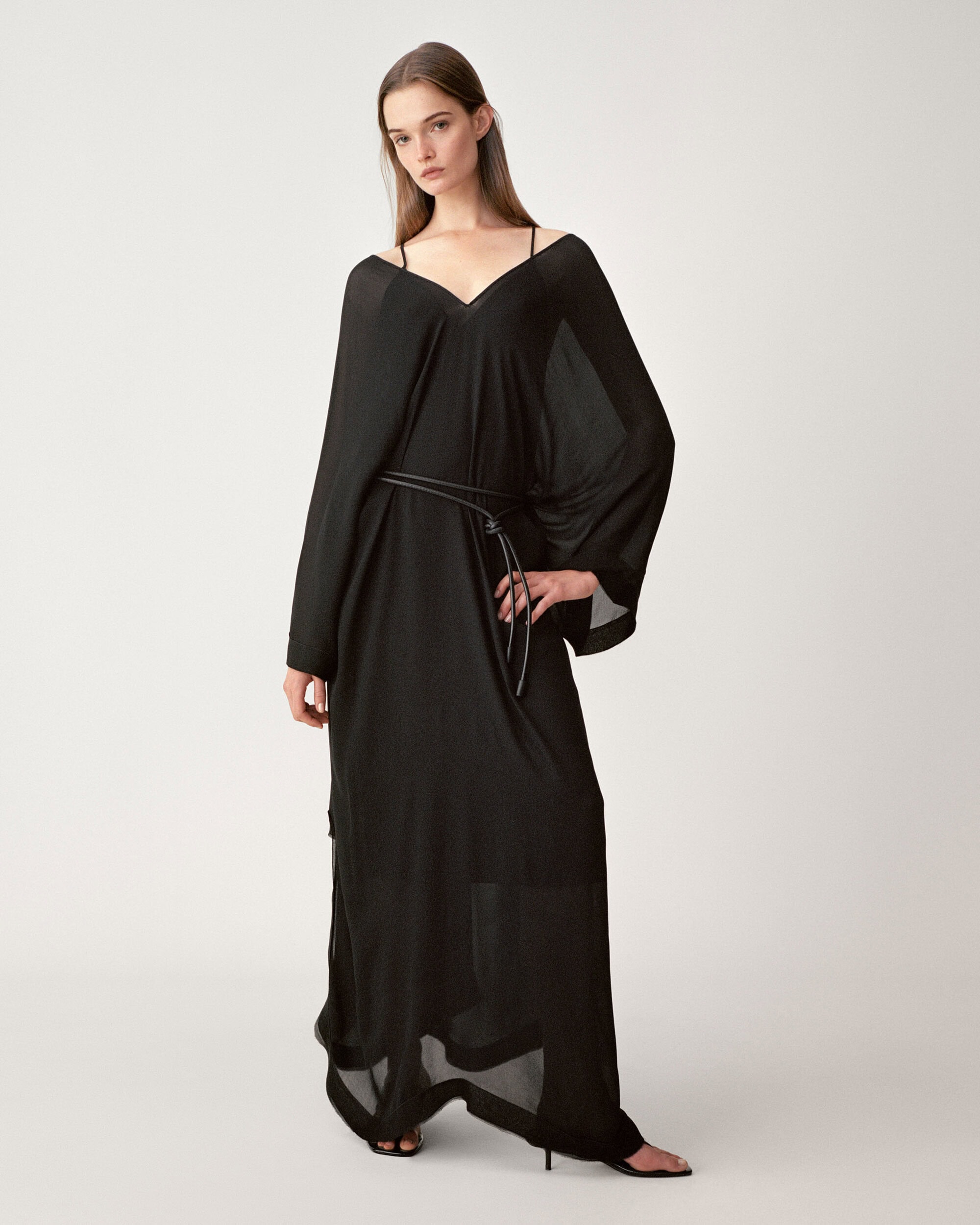 Model wearing a long black sheer dress with a thin belt standing against a seamless white studio background