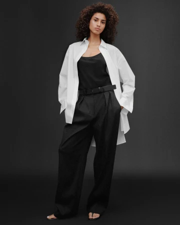 A woman with voluminous dark curly hair wears a white button-down shirt over a black top and trousers against a dark backdrop.